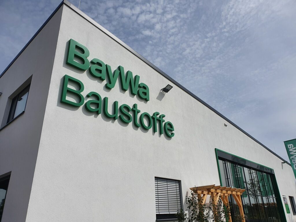 Das Bild zeigt die Außenansicht eines Gebäudes mit der Aufschrift „BayWa Baustoffe“ in großen grünen Buchstaben an der Seite. Das Gebäude hat ein modernes Design mit weißen Wänden und großen Fenstern. Vor dem Eingang befindet sich eine kleine Holzstruktur, möglicherweise eine Pergola, und einige Pflanzen sind vor dem Gebäude sichtbar. Der Himmel ist teilweise bewölkt.