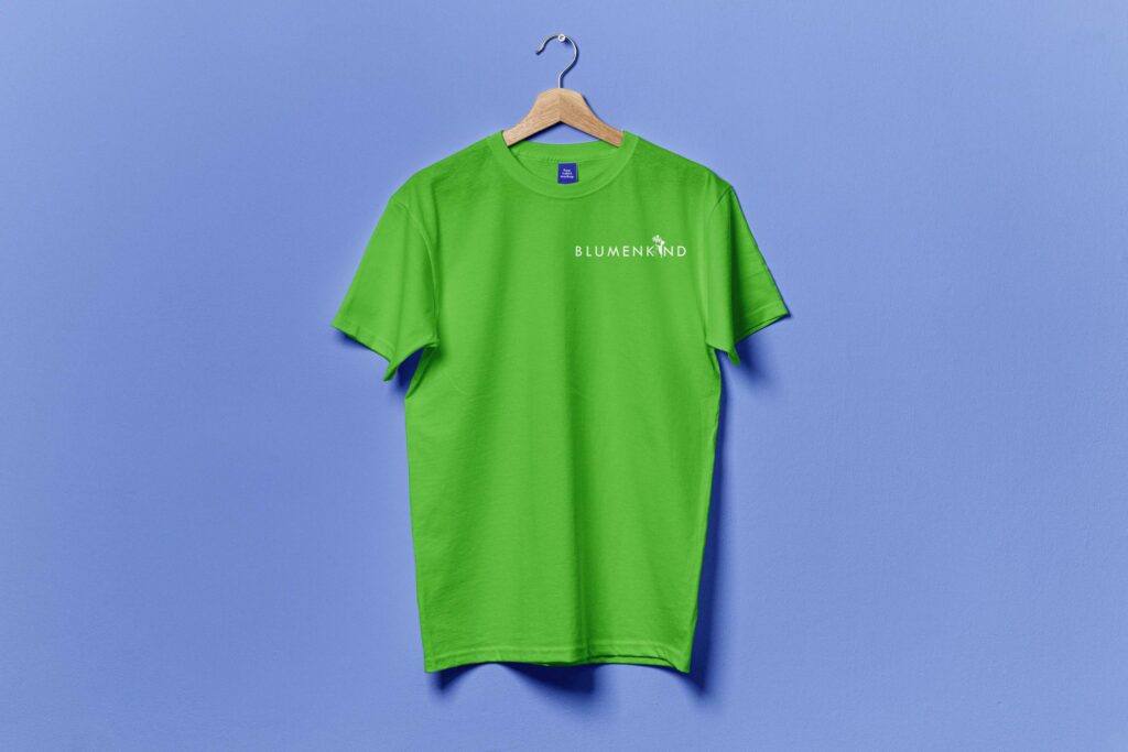 Das Bild zeigt ein grünes T-Shirt auf einem Holz-Kleiderbügel vor einem blauen Hintergrund. Auf dem T-Shirt ist das Logo der Firma "Blumenkind" in weiß bedruckt.