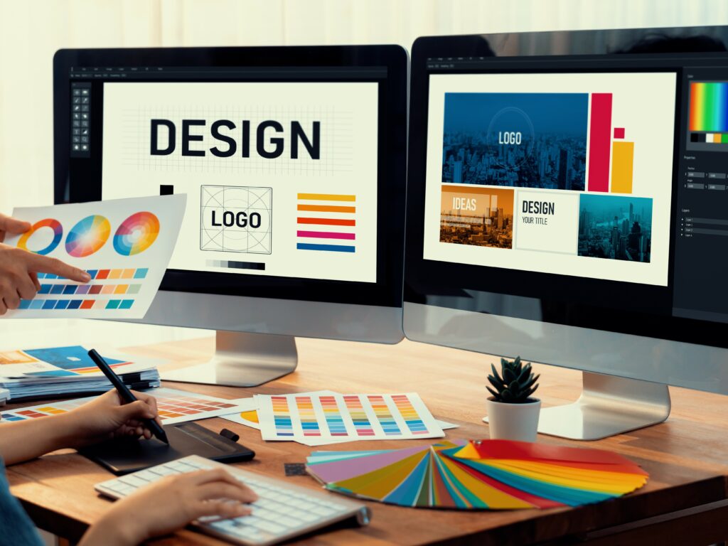 Bildschirme, auf welchen Grafikprogramme geöffnet sind. Auf den Schreibtisch sieht man Farbfächer.